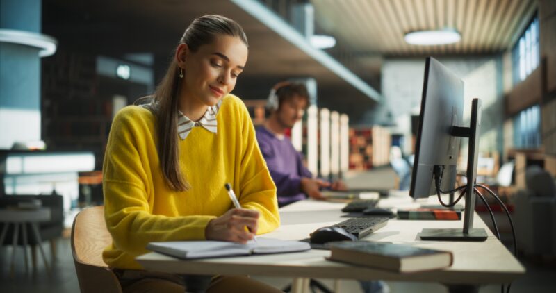 Junge Frau in gelbem Pullover schreibt konzentriert an einem Schreibtisch in moderner Bibliothek oder Lernumgebung – symbolisiert Studium, Prüfungsvorbereitung oder wissenschaftliches Arbeiten.
